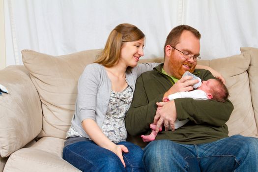 A mother and father hold their newborn baby son in their arms and look towards him showing happiness and caring emotions while bottle feeding the boy.