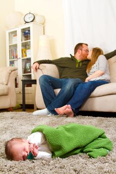 A newborn baby boy sleeps on a comfortable rug while wrapped up in a green blanket with his parents in the background together.