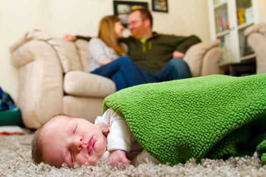 A newborn baby boy sleeps on a comfortable rug while wrapped up in a green blanket with his parents in the background together.