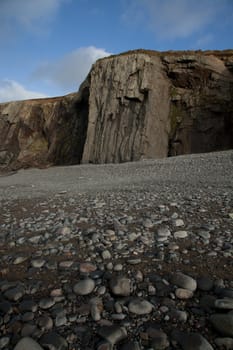 A pebble shore leads to a shale buttress on a cliff with blue sky and cloud in the distance.