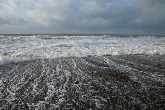 A pebble beach with waves from the sea breaking on the shore with a cloudy sky in the distance.