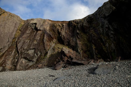 A pebble shore leads to a rock fall under a shale cliff with a blue sky and cloud in the distance.