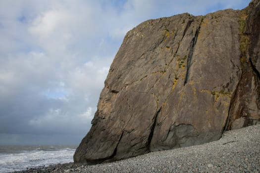 A shale buttress sea cliff with a pebble shore and sea with a cloudy sky overhead.
