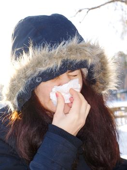 Young woman wearing furry hood, sneezes, holding tissue next to her nose