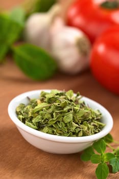 Dried oregano leaves in small bowl with fresh oregano on the side, tomato, garlic and basil in the back (Selective Focus, Focus one third into the dried oregano leaves)