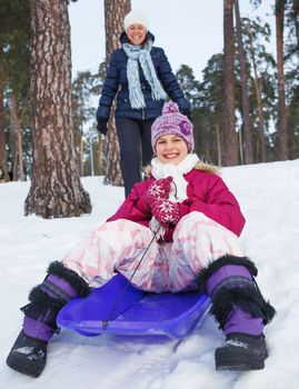 Cute girl on sleds with her mother in snow forest.