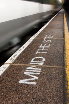 Image of the words, mind the step, painted as a safety warning on a train platform with a train passing.