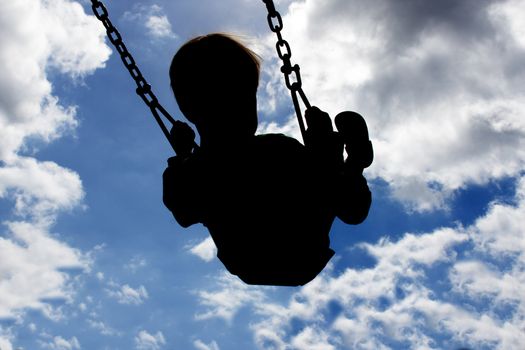 Silloutte of a young child swinging high with a blue sky and white clouds in the background.