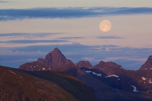 Full moon rising above scenic mountain peaks lit by midnight sun in arctic Europe