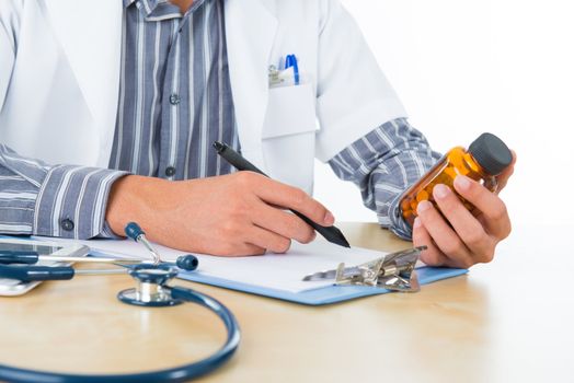 Southeast Asian medical doctor holding a bottle pills and writing on clipboard, sitting in front working desk.