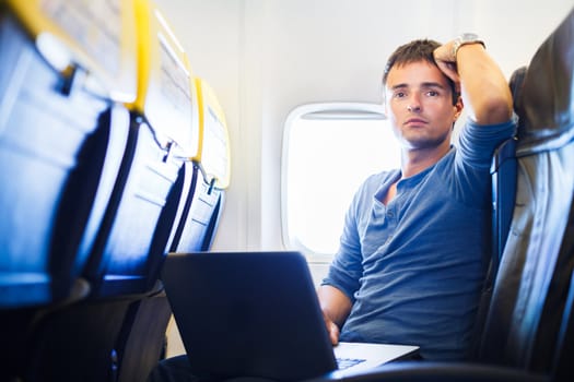 Handsome young man daydreaming while working on his laptop computer on board of an airplane during flight