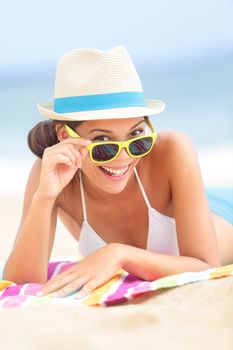 Woman on beach with sunglasses looking flirting at camera smiling happy and joyful during summer vacations holiday travel. Beautiful young multiethnic Asian Chinese / Caucasian hipster lying on beach towel.