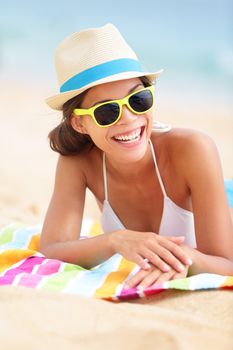 Beach travel woman smiling lying down on beach towel during summer vacation holiday. Mixed race Asian Chinese / Caucasian girl hipster wearing sunglasses and hat.