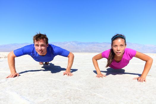 Sport - young fitness couple athletes doing push ups outdoors in desert nature landscape. Caucasian man sports model and Asian woman fitness model doing push-ups exercise under the burning sun.