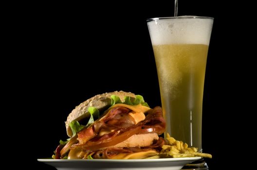 Spilling Glass of Beer and Big Tasty Burger with Fried Bacon, Lettuce, Tomato, Cheese and French Fries isolated on black background