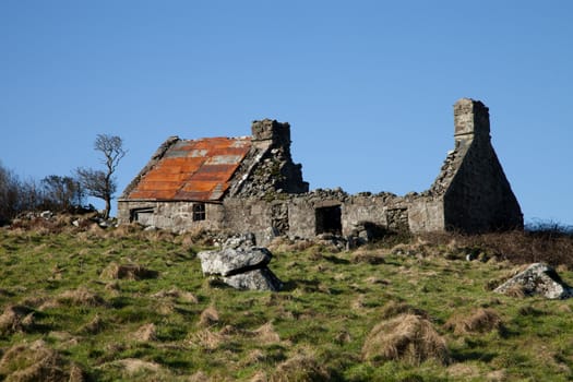 A stone built derelict building with partial tin roof and chimneys in a field.