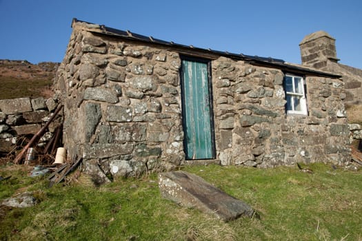 A stone built tool shed with window and door on grass and a blue sky in the background.