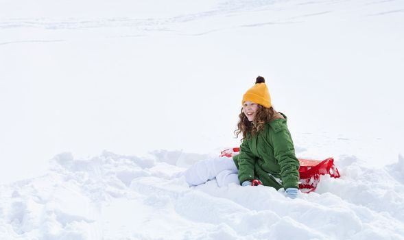 Smiling teenager girl with red plastic sledge sitting in snow 