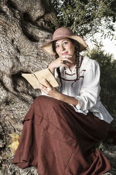 View of a beautiful girl in a classic dress in a countryside set in the shade of a very old olive tree reading a book.