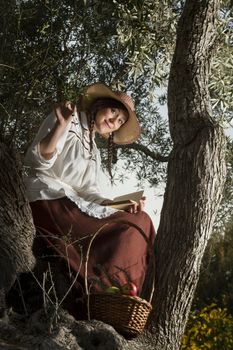 View of a beautiful girl in a classic dress in a countryside set in the shade of a very old olive tree reading a book.