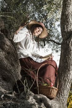 View of a beautiful girl in a classic dress in a countryside set in the shade of a very old olive tree reading a book.