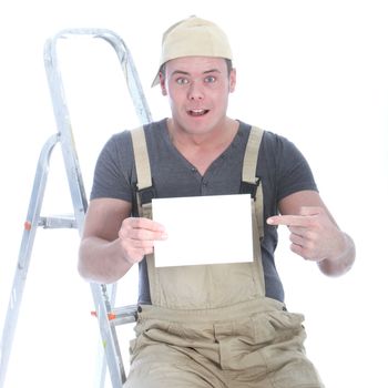 Shocked handsome young handyman balanced on his ladder pointing to a blank white sign with copyspace for your text that he is holding in his hand