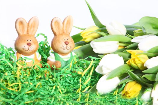 Two cute little Easter bunnies peering out of green straw with a large bunch of fresh spring tulips in yellow and white alongside them