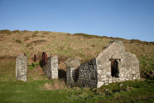 Historic industrial, mine buildings built of stone with a rusty turning wheel infront of a slope with a blue sky in the distance.