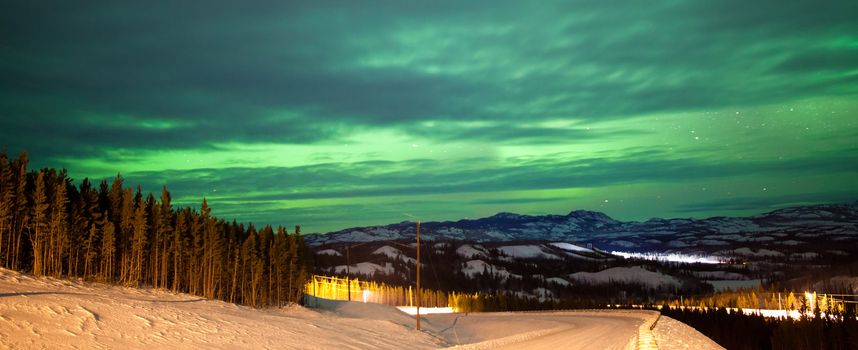 Northern Lights or Aurora borealis or polar lights illuminating overcast sky over snowy winter road landscape