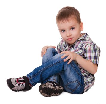Small serious sitting boy in jeans and sneakers isolated on a white background 