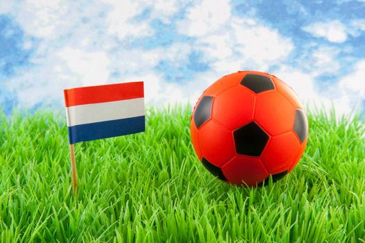 Orange ball and Dutch flag on soccer field against paper blue cloudy sky 
