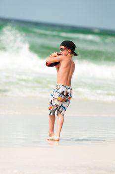 Young boy in Swim trunks playing football at the beach with the ocean in the background.