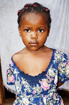 Portrait of a cute and sweet little black African girl, smiling but looking a bit shy, posing in front of her house, great details.