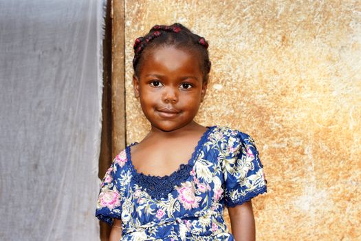 Portrait of a cute and sweet little black African girl, smiling but looking a bit shy, posing in front of her house.