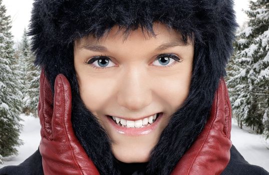 Winter time: portrait of smiling young woman outside in the snowy forest where it is very cold, keeping warm with her black faux fur hat and red leather gloves.