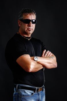 Somber tough guy with tatoos and black sunglasses, arms crossed, looking at camera, studio shot over black.
