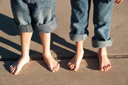 Two children with bare feet and  pants rolled up standing on sidewalk.