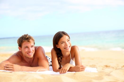 Couple on beach looking happy during summer travel vacation holidays. Multiracial young couple lying in sand on beach on looking to side. Asian woman, Caucasian man.