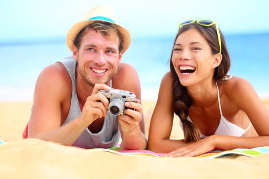 Young happy multicultural couple on beach having fun laughing during summer holiday vacation. Caucasian hipster man with retro vintage camera and asian woman joyful.