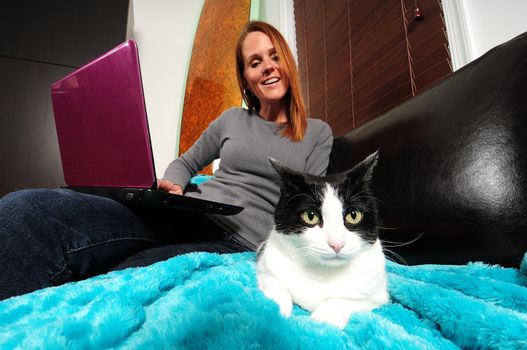 Pretty young woman smiling with black and white cat while sitting on the couch with computer
