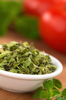 Dried oregano leaves in small bowl with fresh oregano on the side, tomato and basil in the back (Selective Focus, Focus one third into the dried oregano leaves)