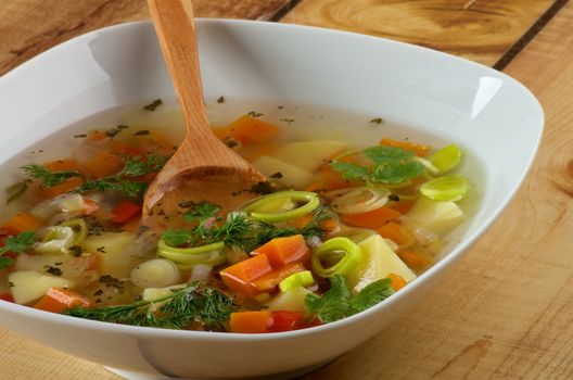 White Bowl of Vegetable Soup with Potato, Carrot, Bell Pepper, Leek, Parsley and Dill with Wooden Spoon closeup on Wooden background