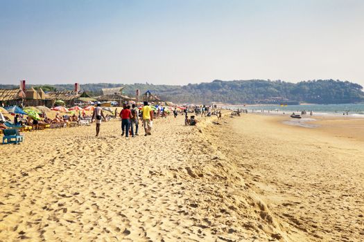Horizontal capture of a group of young men strolling on a busy sandy beach full of holiday makers on a sunny afternoon. Location of this landscape was Goa, India