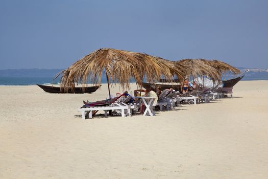 Landscape of secluded beach with day trippers and holidaymakers basking in the sun under the shade of coconut parasols. White sand, blue sky, sea, surf and fishing boats for a backdrop. Generic tropica shot location, Goa India