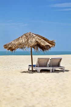 A generic vertical portrait of a tropical deserted beach with loungers under a coconut leaf parasol. Blue sky, ocean, gentle surf sound and clean sandy beach. Location, Goa India