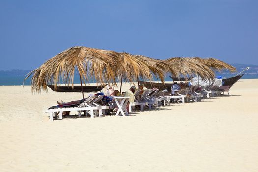 Landscape of secluded beach and a row of tropical parasols with day trippers and holidaymakers basking in the sun under the shade. White sand, blue sky, sea, surf and fishing boats for a backdrop. Generic tropical shot location, Goa India