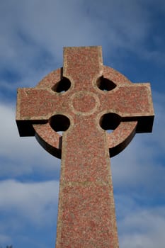 A polished red stone carved celtic style christian cross against a blue sky with cloud.