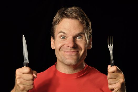 Closeup of hungry Caucasian man in red shirt holding knife and fork on black background while making silly face