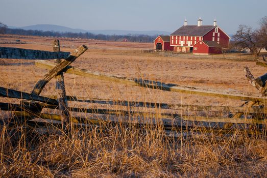 Photograph of a 4 rail fencing system dating back to the civil war era.  Large red barn in the distance and fallow field in the foreground.