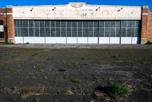 Photograph of an abandoned Aircraft Hangar with faded paint and text.  Weeds are growing up though the old tarmac.